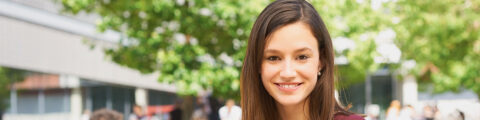 Das Bild zeigt eine lächelnde junge Frau mit schulterlangen Haaren, die direkt in die Kamera blickt. Im Hintergrund sind verschwommen mehrere Personen und ein Baum zu sehen.